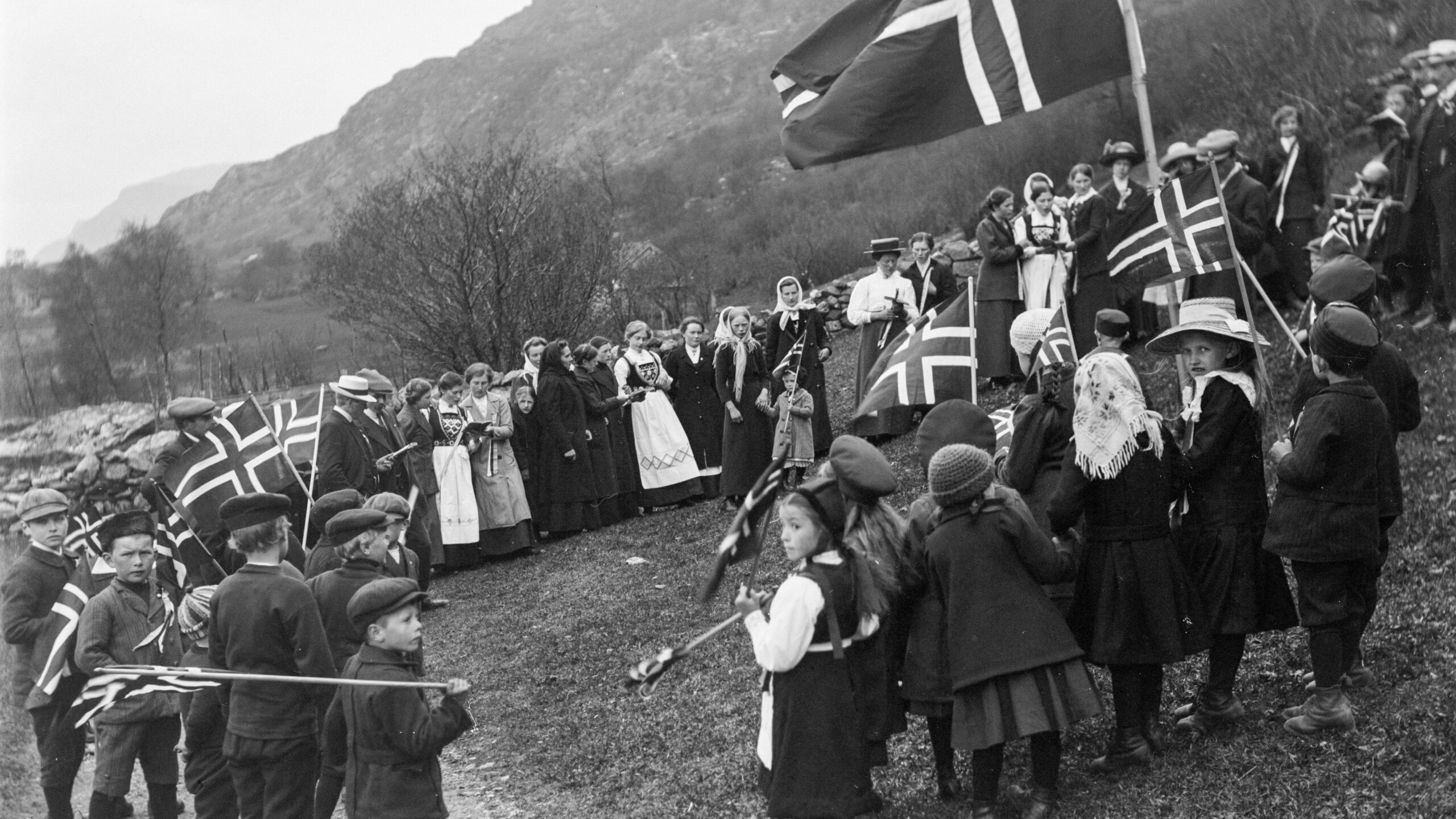 Sort.hvitt bilde fra 17. mai, tidlig 1900-tallet. Mange barn og voksne er samlet i en skråning. De holder flagg og er pent kledd. Noen har bunad.