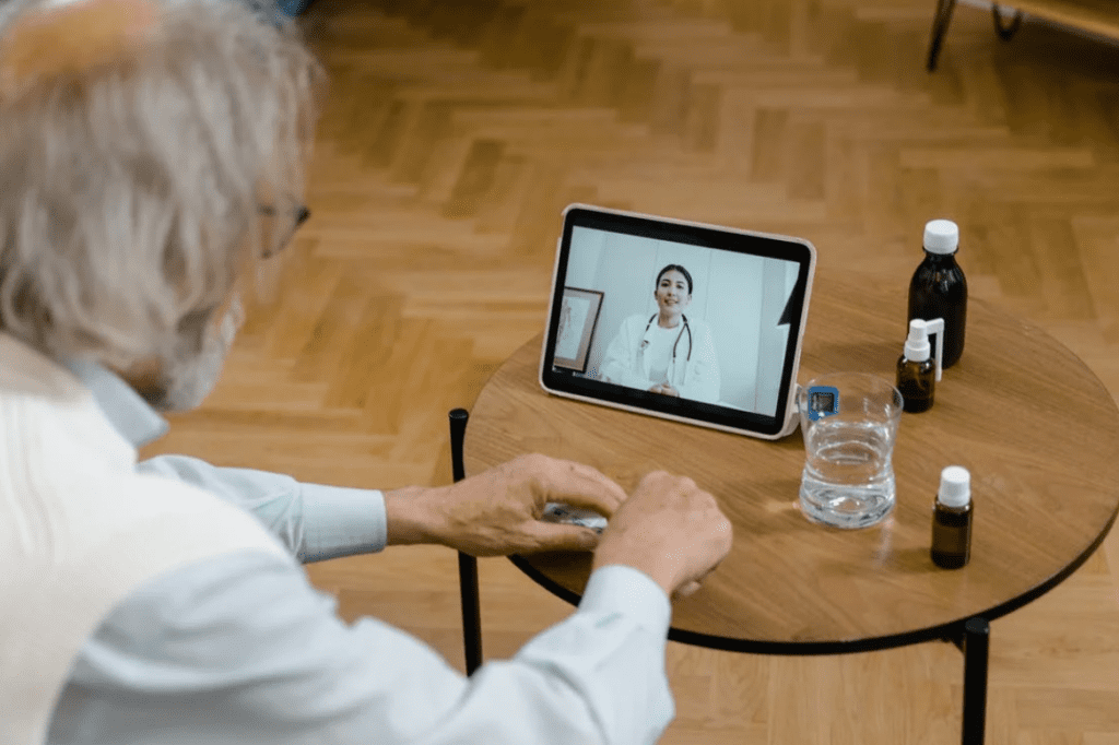 Eldre mann grått hår sitter foran skjerm og snakker med lege
