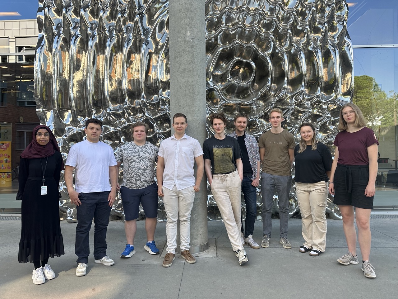 En gruppe med unge mennesker står stilt mot en kunst installasjon og smiler mot kameraet.