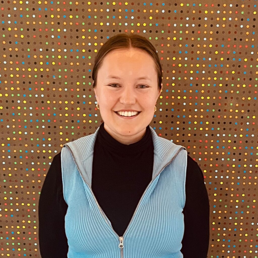 Sofie står mot en prikkete bakgrunn. Hun ser rett mot kameraet og smiler. Hun har håret opp, lyseblå vest og sort langermet genser.