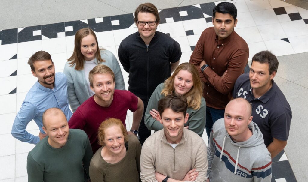 En gruppe unge mennesker er fotografert ovenfra. Det er en professjonell setting og de ser mot kameraet og smiler. Det er elleve mennesker avbildet. 