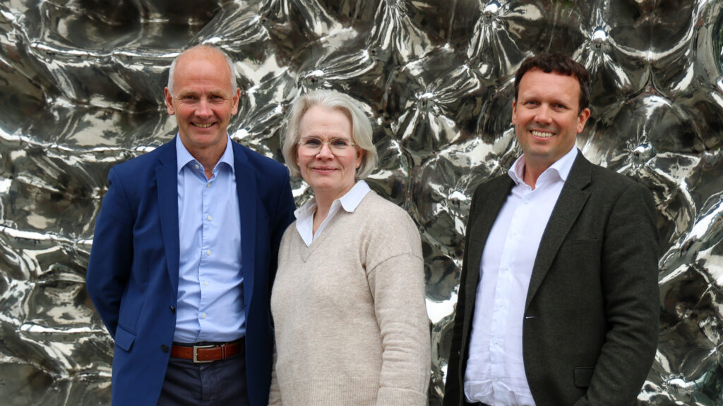 Bildet viser tre mennesker smilende mot kameraet. I bakgrunnen er en bølgete vegg i metall som ligner på et speil. Dette er en professjonell setting.