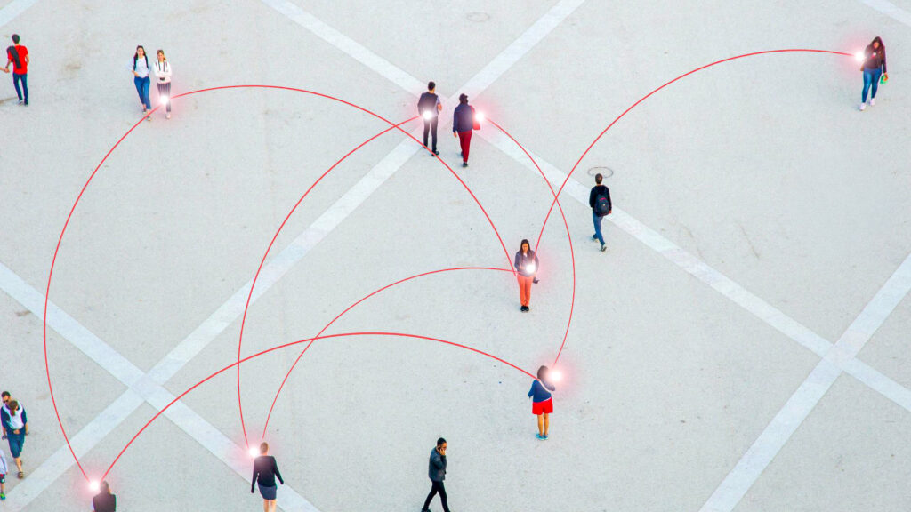 Bildet viser en spredt gruppe med mennesker som går over en betongflate. Det er røde, kurvede linjer mellpom enkelte av menneskene for å illustrere smittespredning. Flybilde.
