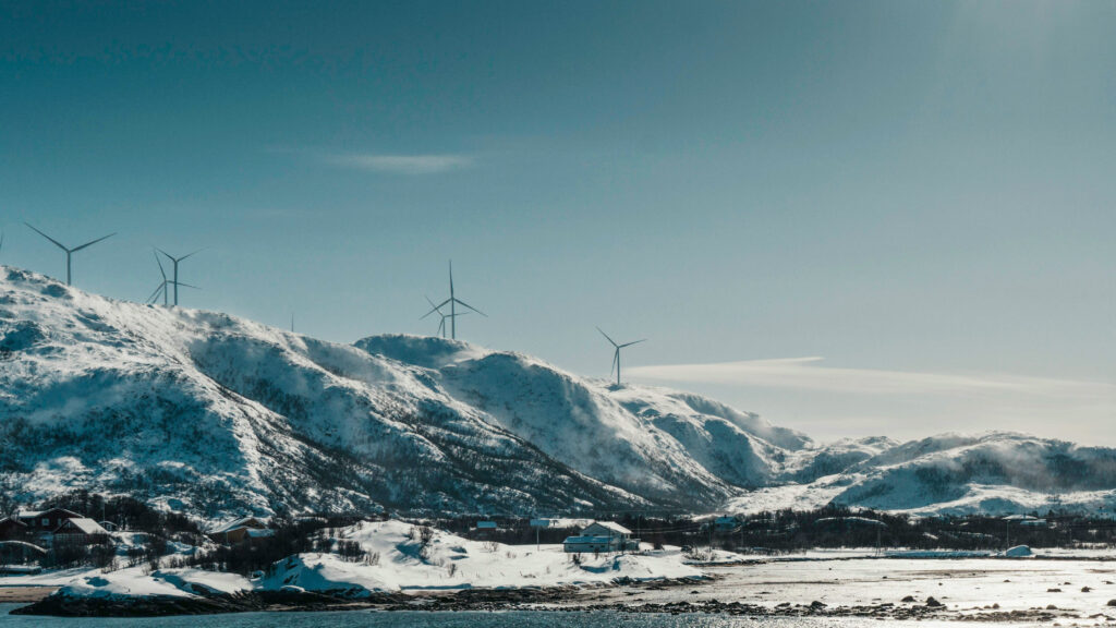 Landscape image of snow-covered mountains with windmills on top of the peaks. Some buildings scattered around the landscape. Sunny day and blue skies.