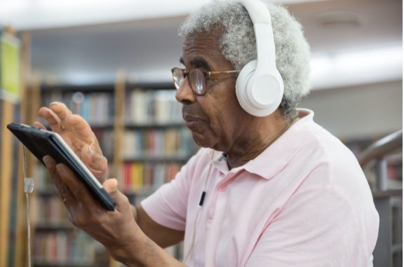 An elderly man wearing glasses and headphones is using a digital tablet.