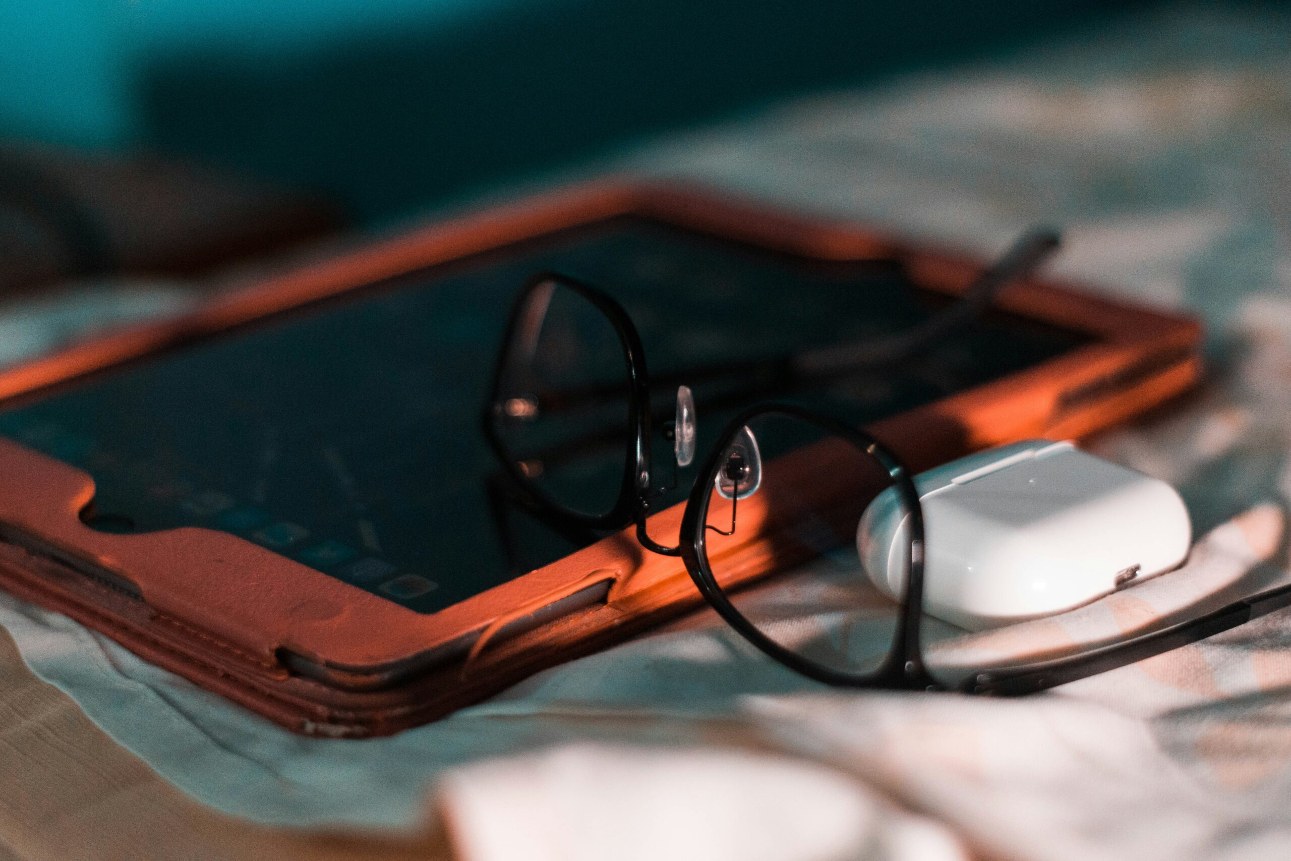 The image features an ipad in a red case, black-rimmed glasses and an airpod case.