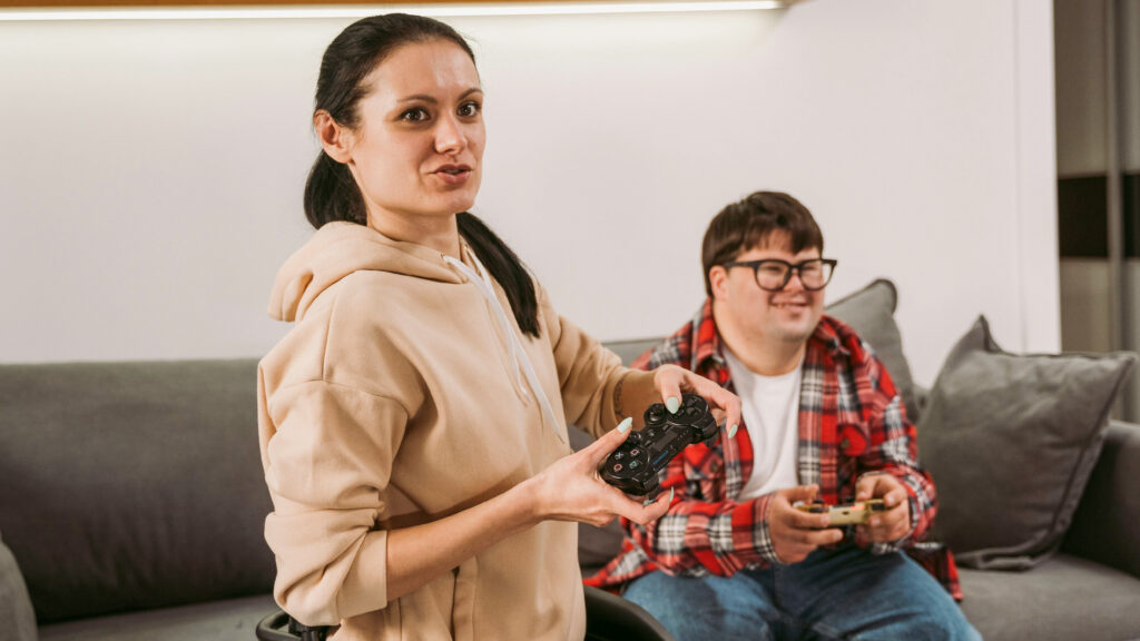 Two individuals are playing video games together, focused on a screen outside the frame. On the left is a woman in a wheelchair, wearing a beige hoodie and black pants, with long hair gathered over her left shoulder. On the right is a man seated on the couch, wearing glasses, a red plaid shirt over a white t-shirt, and blue jeans. He appears to have Down syndrome. Both are holding game controllers and seem to be having fun while being engaged in the game.