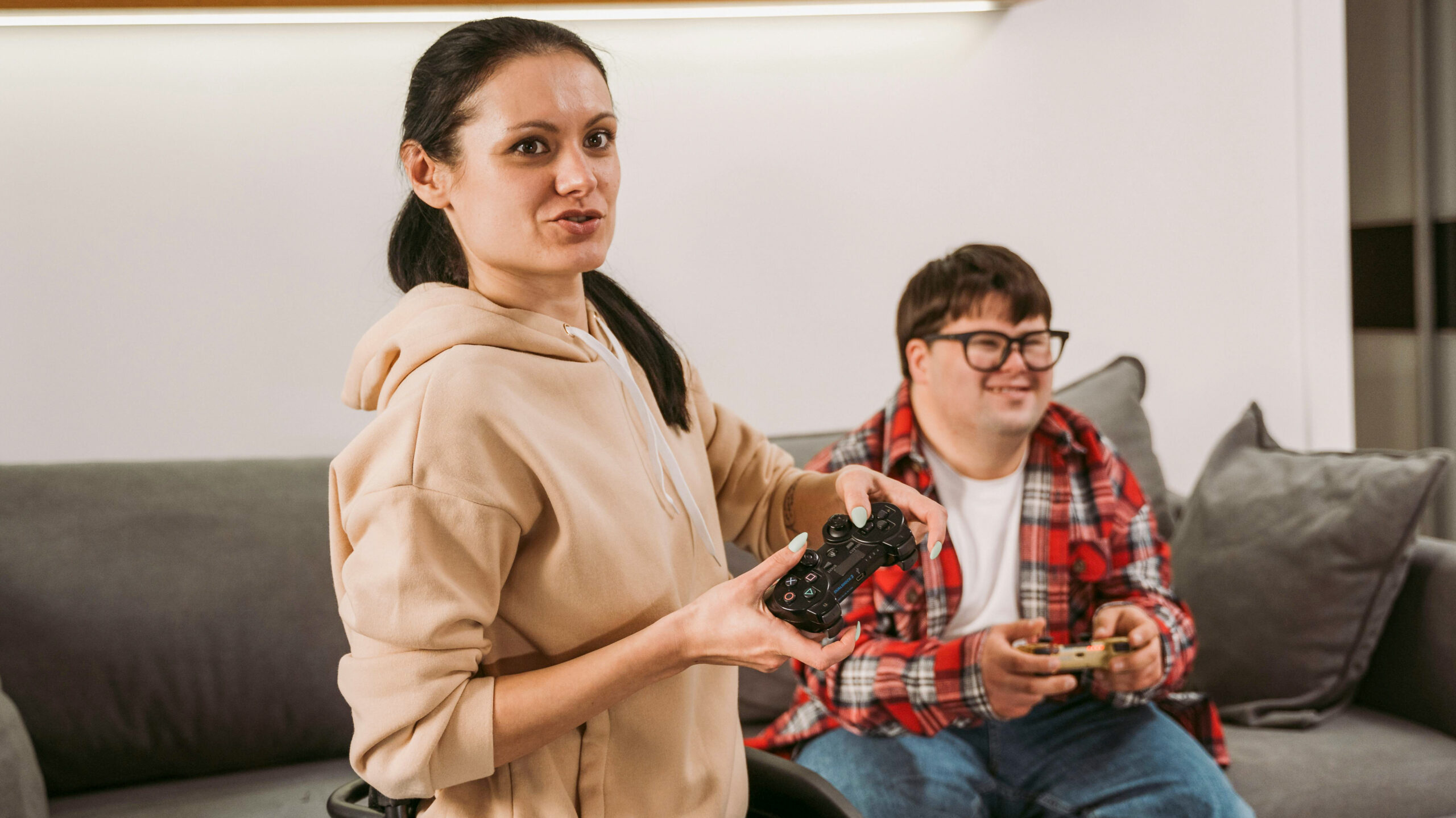Two individuals are playing video games together, focused on a screen outside the frame. On the left is a woman in a wheelchair, wearing a beige hoodie and black pants, with long hair gathered over her left shoulder. On the right is a man seated on the couch, wearing glasses, a red plaid shirt over a white t-shirt, and blue jeans. He appears to have Down syndrome. Both are holding game controllers and seem to be having fun while being engaged in the game.