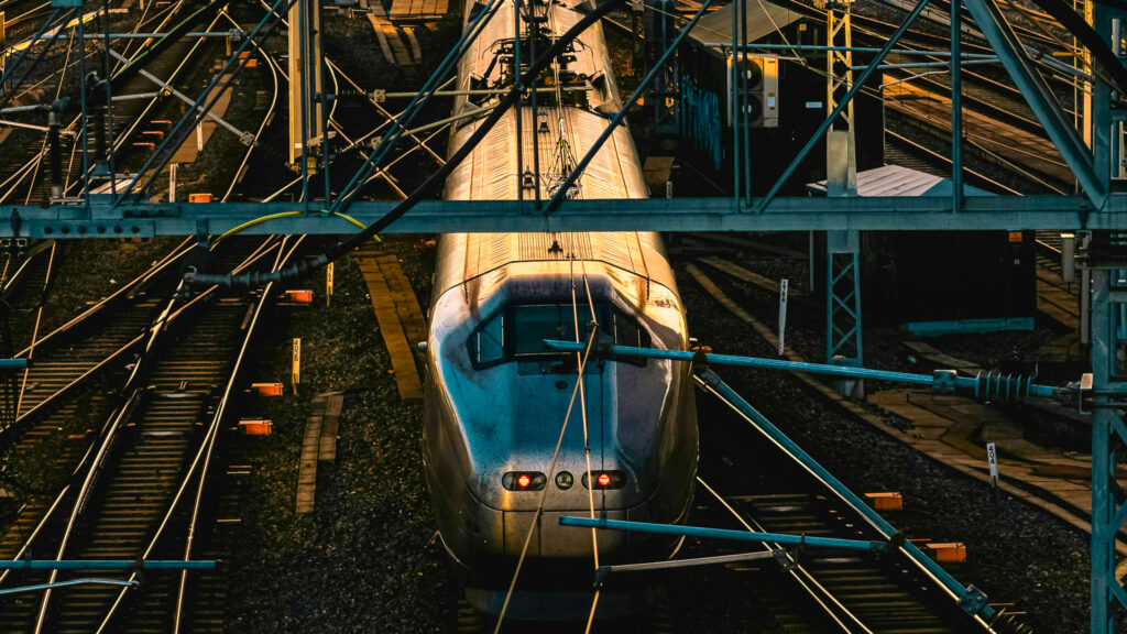 The image shows a train on the tracks with several overhead power lines and cables above it.