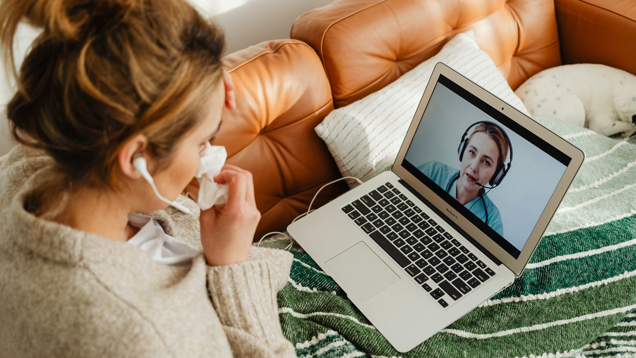 A young woman sits on the sofa with a laptop on her laptop. On the screen is a healthcare professional with a headset on.